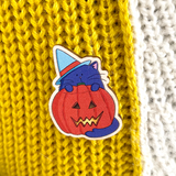 A wooden pin badge featuring a purple cat wearing a blue witches hat and standing in front of a lit halloween pumpkin is shown on a wooly jumper